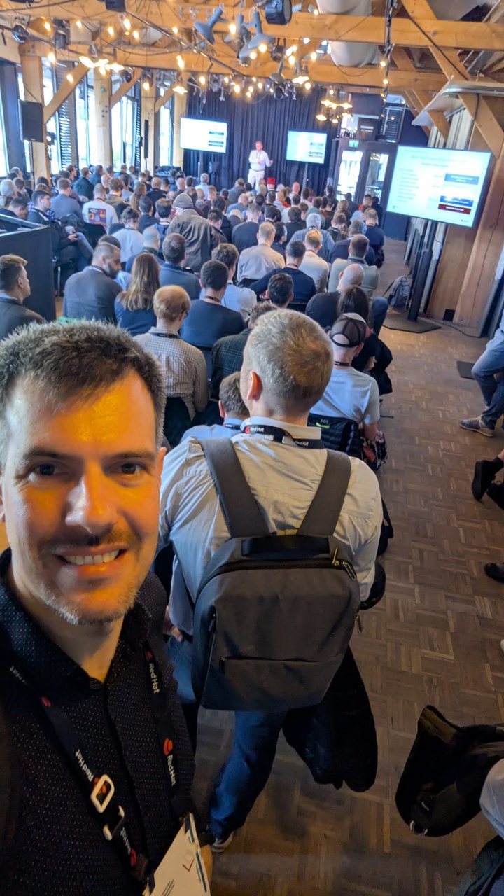 Luca Berton in the side aisle of the packed main hall at OpenShift Commons Gathering Amsterdam 2026, with a speaker on stage and slides on the screens