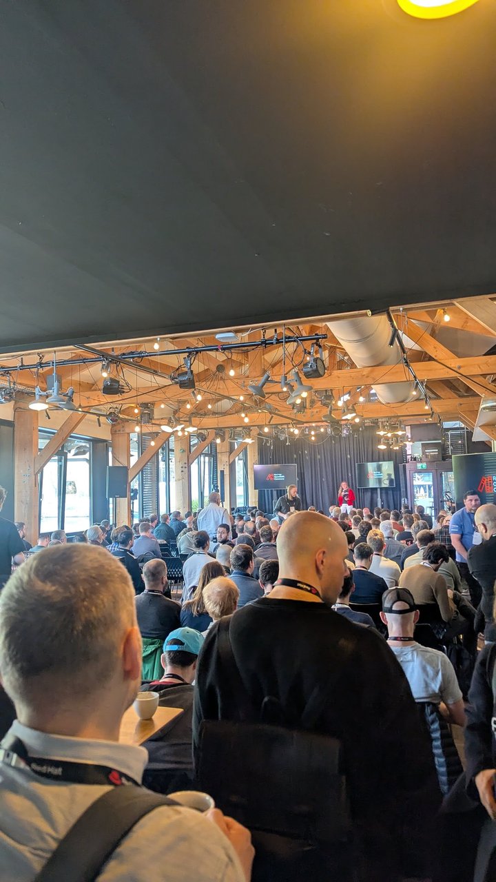 The stage at OpenShift Commons Gathering Amsterdam 2026, with Red Hat OpenShift Commons logos on the screens and a full room under the timber roof