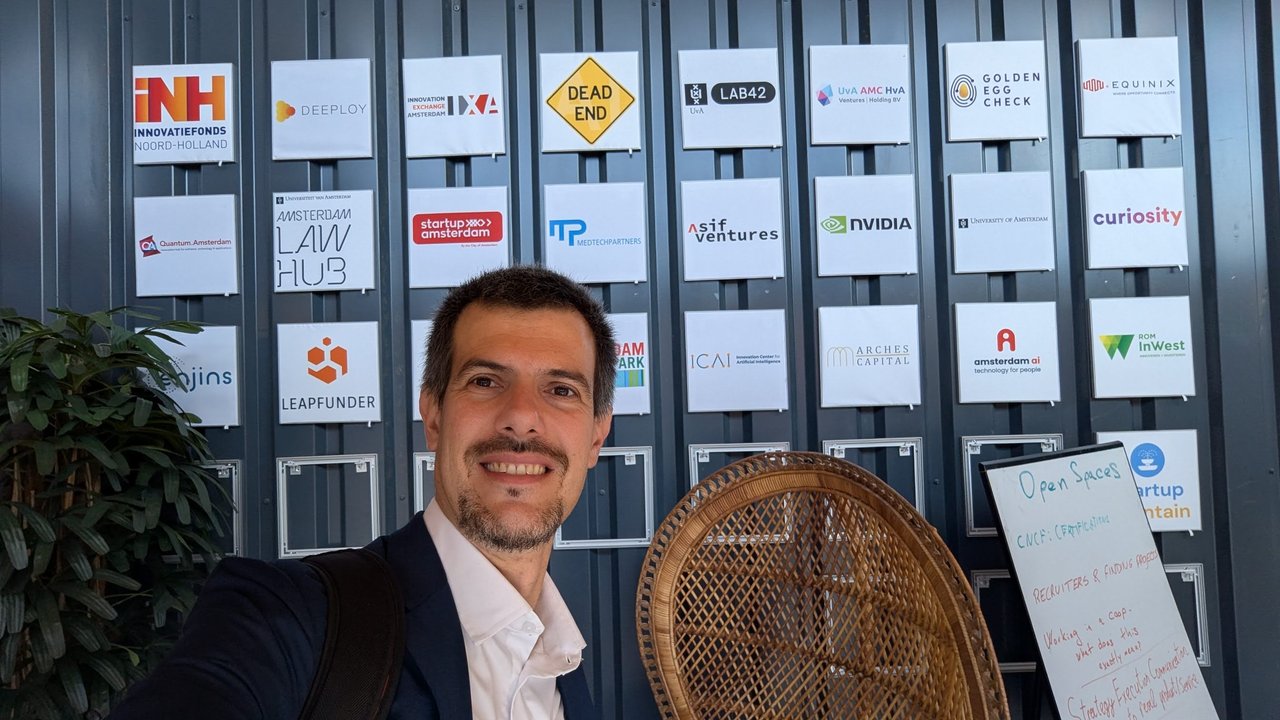 Selfie of Luca Berton in front of a wall of partner logos, next to a whiteboard titled Open Spaces listing topics such as CNCF certifications, recruiters and finding projects, and working in a coop