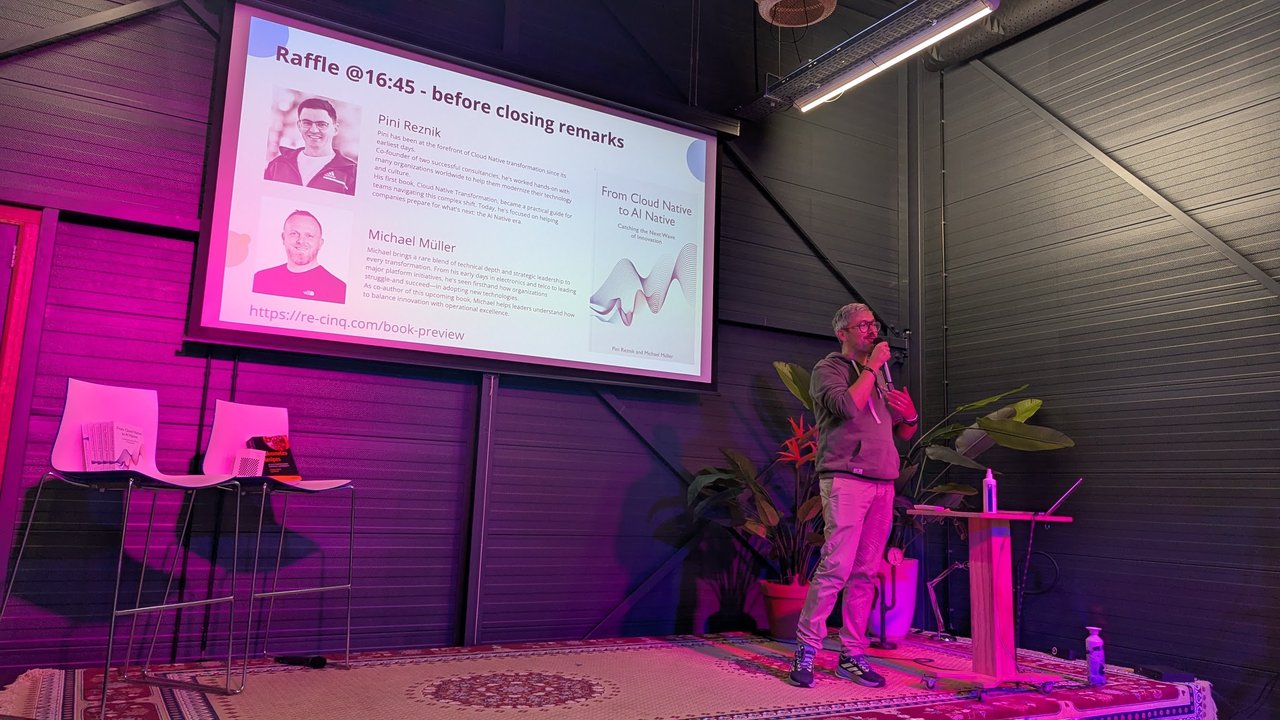FikaWorks Day stage with a slide titled Raffle at 16:45, before closing remarks, introducing Pini Reznik and Michael Müller and the book From Cloud Native to AI Native, while Pini speaks; book copies rest on a stool