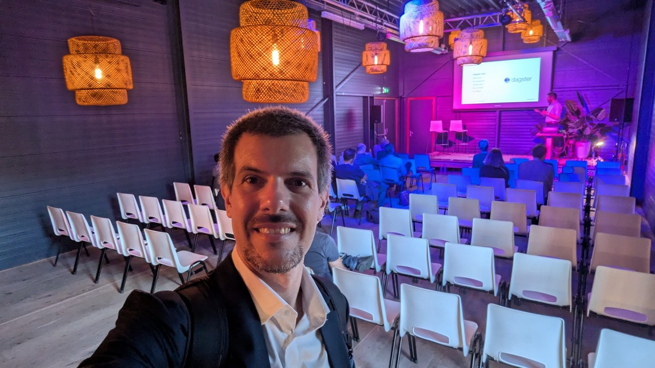 Selfie of Luca Berton in the FikaWorks Day event space, rows of white chairs and a speaker on stage with a Dagster slide on the screen