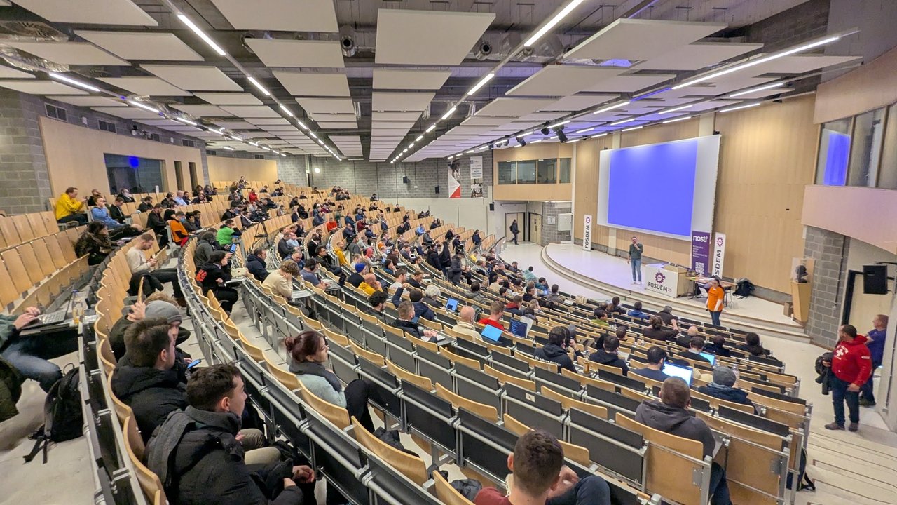 A large FOSDEM 2025 auditorium on Sunday afternoon, with a Nostr banner next to the stage and the audience spread over the tiered seats