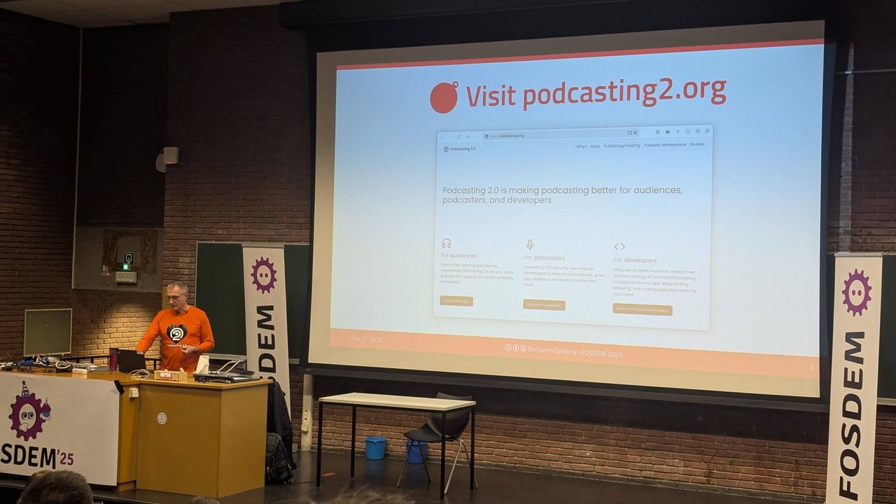 A speaker at the lectern next to a slide saying Visit podcasting2.org, with FOSDEM banners on both sides of the screen