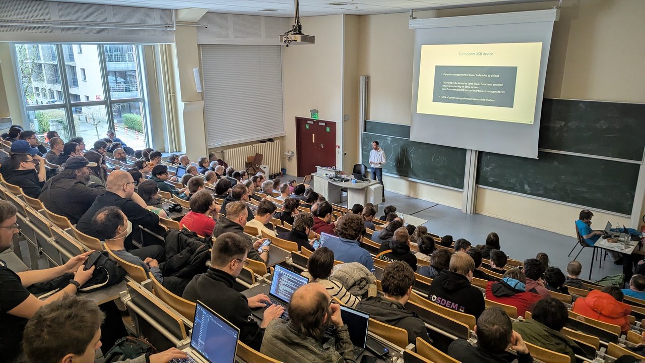 Turn down USB device slide noting 56 percent power saving when turning down a USB modem, in a packed room UA2.114 at FOSDEM 2026