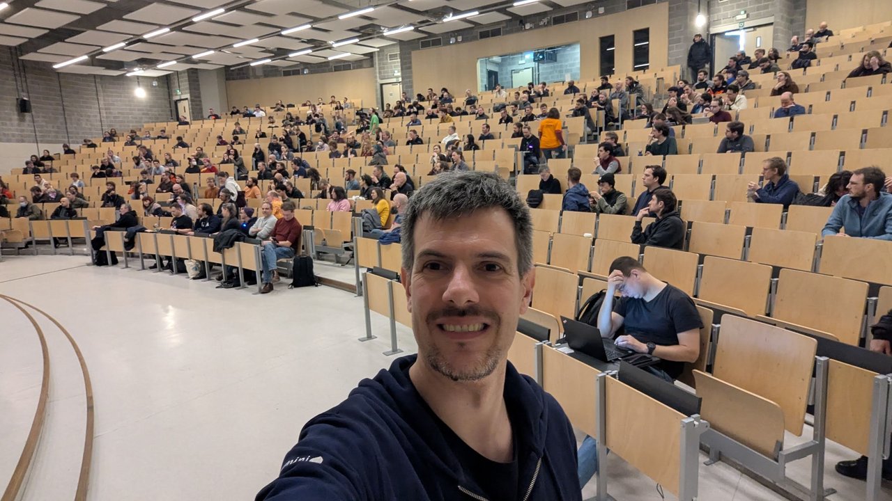 Luca Berton taking a selfie in a large, half-full auditorium at FOSDEM 2026 on Sunday