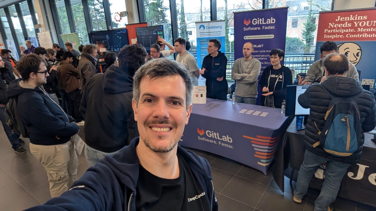 Luca Berton taking a selfie at the GitLab stand at FOSDEM 2026 on Sunday, with visitors at the tables behind him