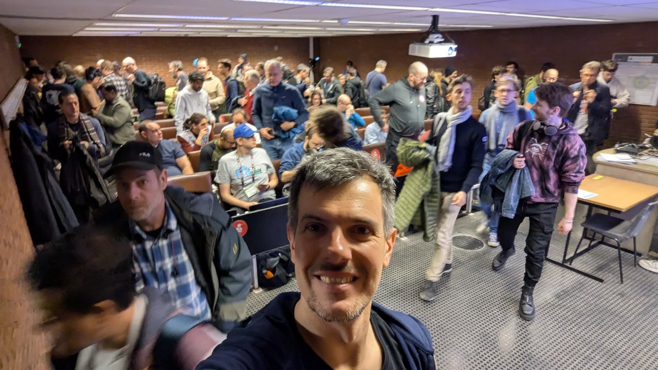 Luca Berton taking a selfie in a crowded FOSDEM 2026 devroom on Saturday, with attendees moving between rows of seats