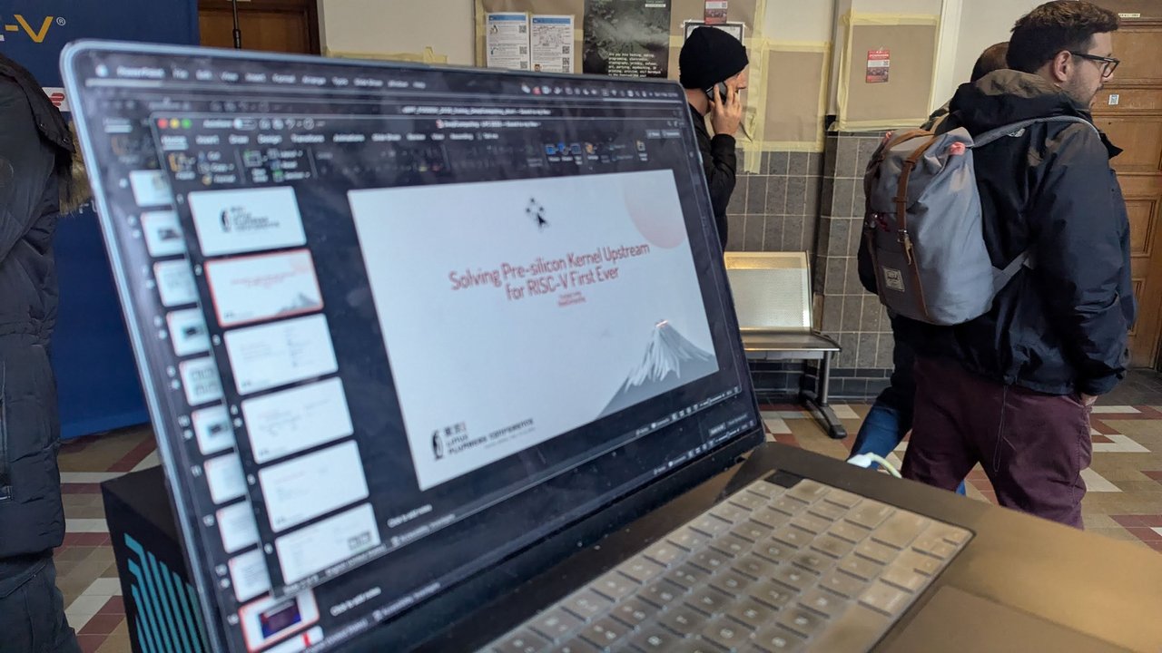 A laptop on a FOSDEM 2026 stand showing a slide deck titled Solving Pre-silicon Kernel Upstream for RISC-V First Ever