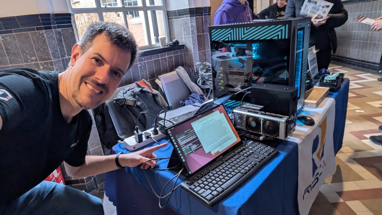 Luca Berton at the RISC-V stand at FOSDEM 2026, pointing at a laptop on a table next to a large tower PC with a glass side panel