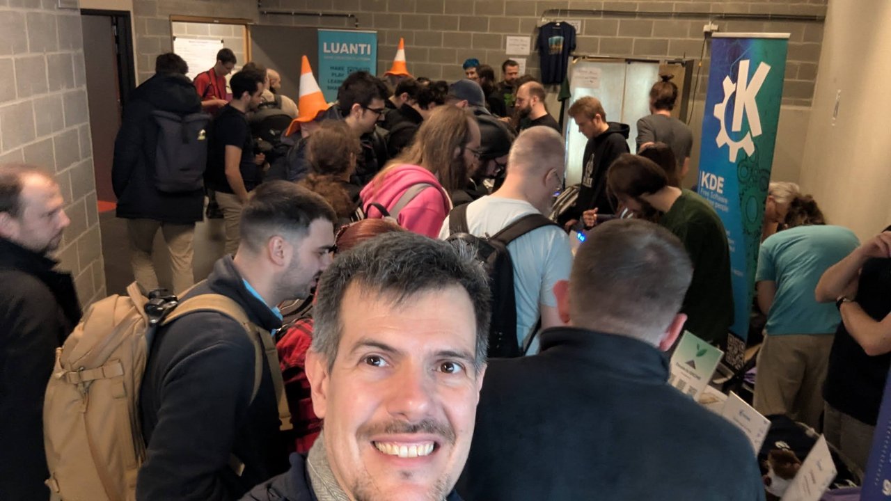 Luca Berton taking a selfie in a crowded corridor of FOSDEM 2026 stands, with KDE and Luanti banners behind him