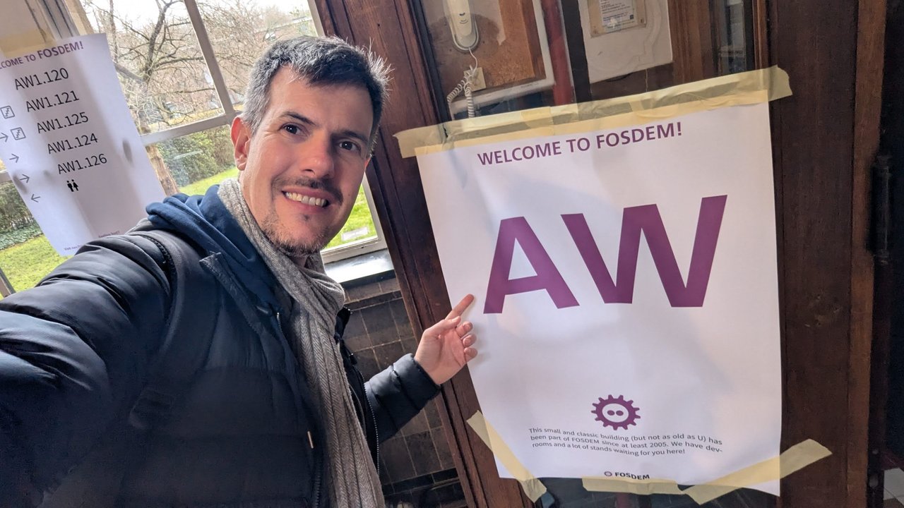 Luca Berton pointing at the Welcome to FOSDEM AW sign on the AW building door, listing rooms AW1.120 to AW1.126, at FOSDEM 2026