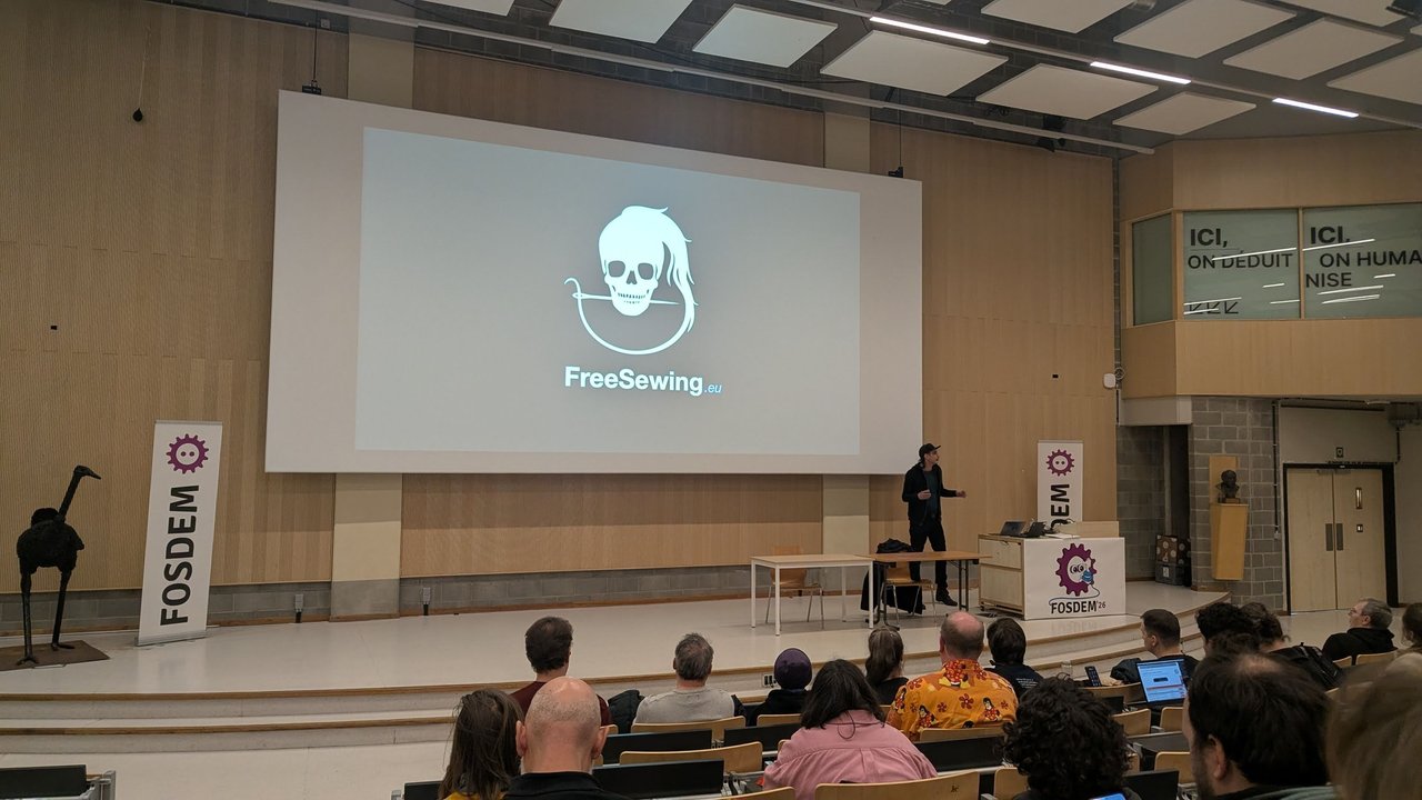 FreeSewing.eu logo on the big screen in a large FOSDEM 2026 auditorium on Sunday, with FOSDEM banners beside the stage