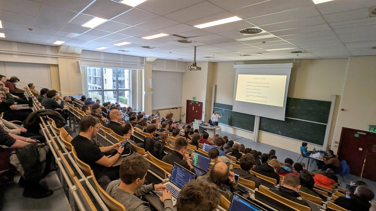 A packed tiered devroom, UA2.114, during a Linux power-saving talk at FOSDEM 2026 on Sunday afternoon