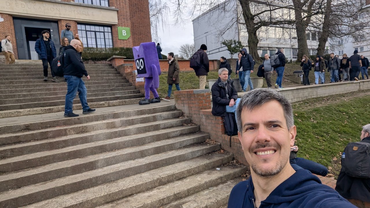 Luca Berton on the ULB campus steps outside building B at FOSDEM 2026, with attendees and a purple mascot on the stairs