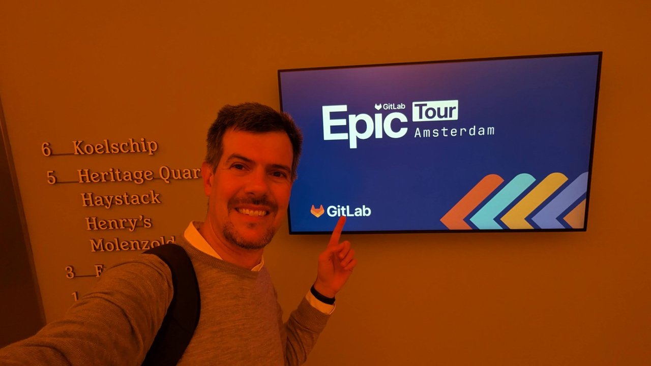 Luca Berton pointing at a GitLab Epic Tour Amsterdam screen in a hallway, with a list of Heineken Experience room names on the wall
