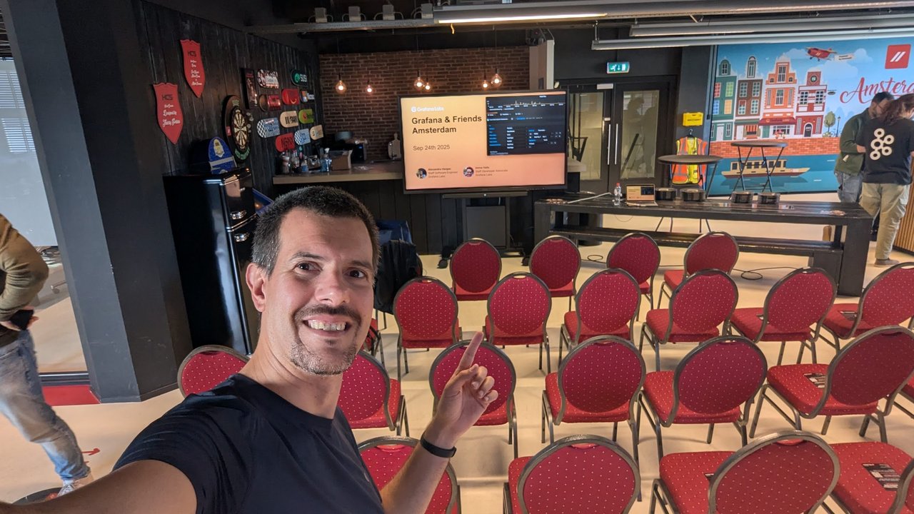 Luca Berton pointing at the Grafana & Friends Amsterdam, Sep 24th 2025 title slide, with empty red chairs and the Amsterdam mural on the right
