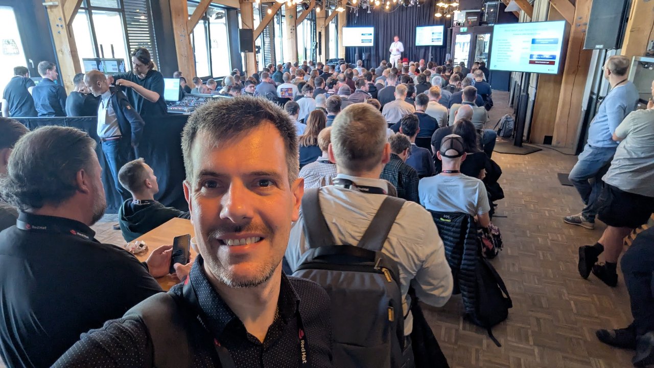 Luca Berton in the packed main hall at OpenShift Commons Gathering Amsterdam 2026, with a speaker on stage and slides on the screens along the room