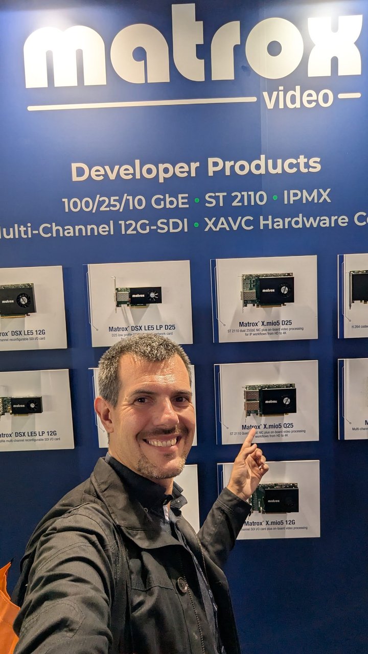 Luca Berton pointing at the Matrox X.mio5 Q25 card on the Matrox Video developer products wall, under the line 100/25/10 GbE, ST 2110, IPMX, Multi-Channel 12G-SDI
