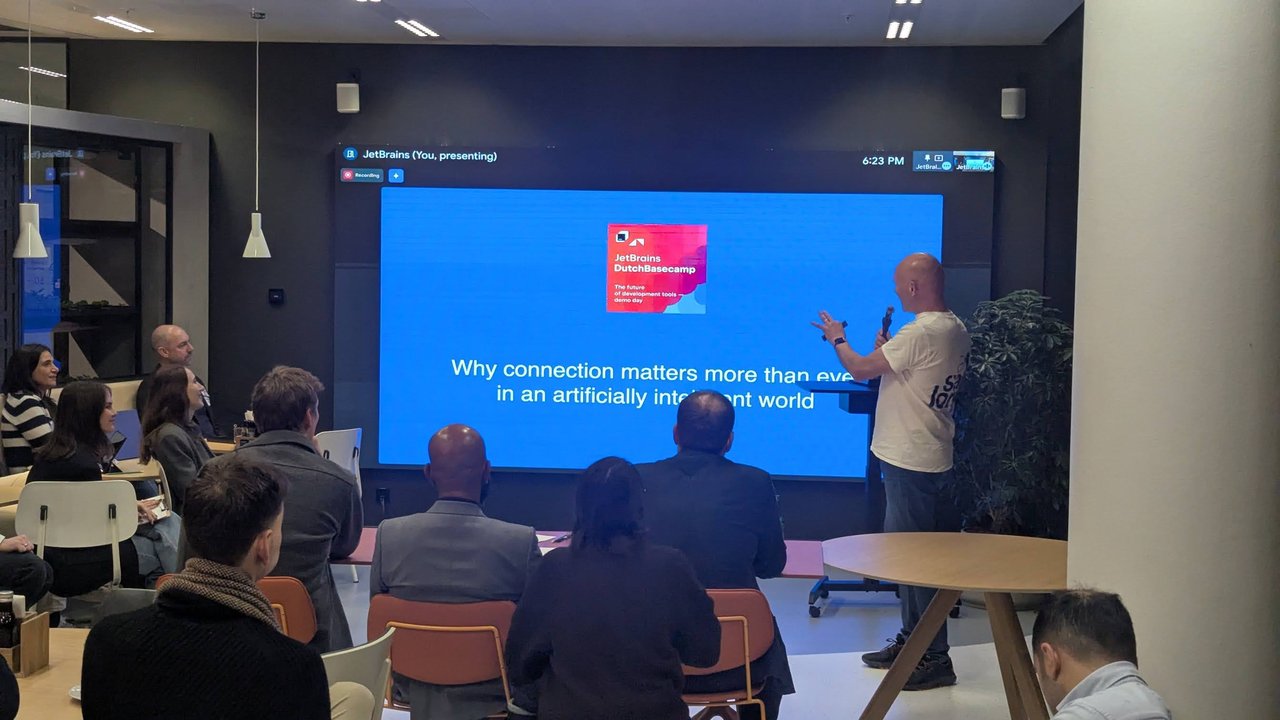 The keynote speaker on stage in front of a blue slide reading Why connection matters more than ever in an artificially intelligent world, with the JetBrains Dutch Basecamp demo day card above it
