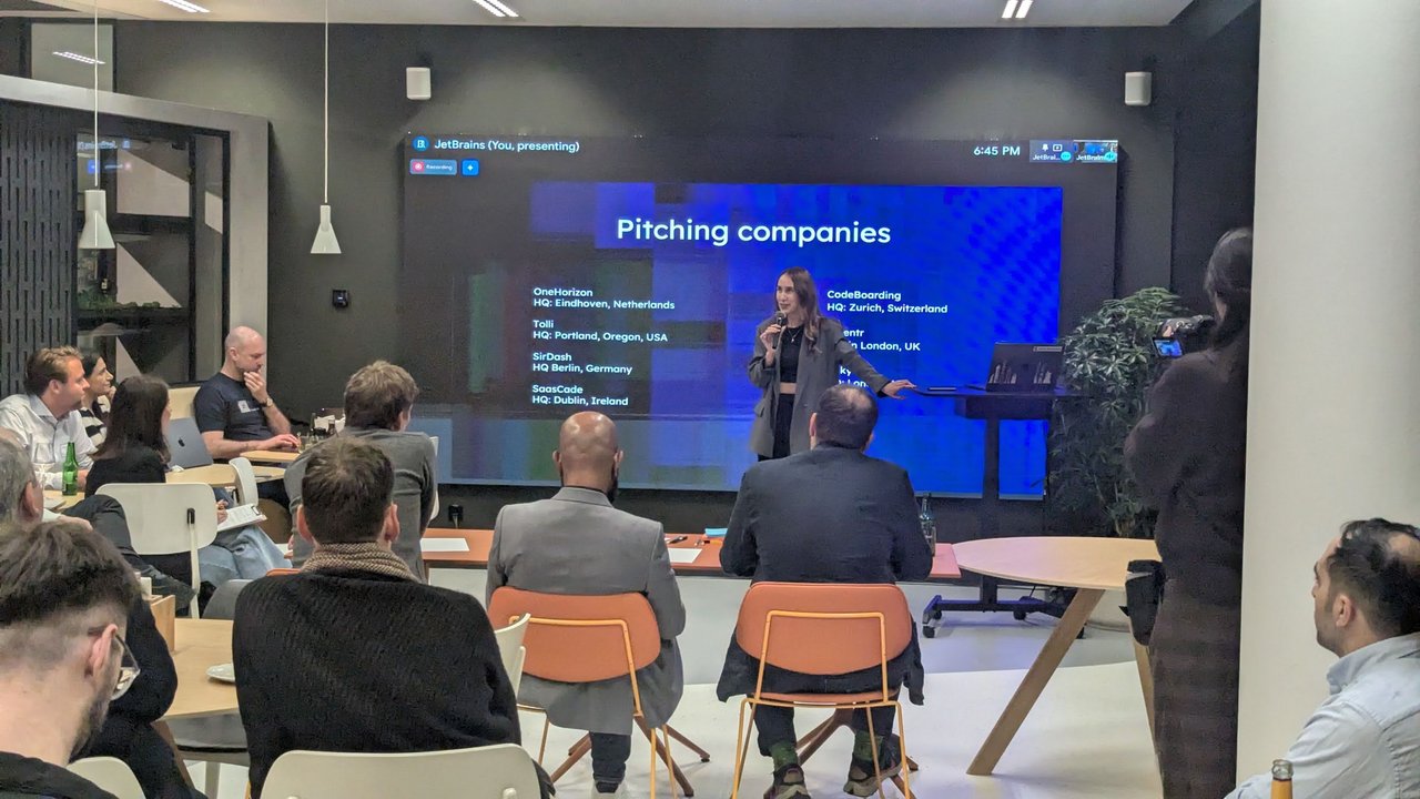 The Pitching companies slide listing OneHorizon in Eindhoven, Tolli in Portland, SirDash in Berlin, SaasCade in Dublin, CodeBoarding in Zurich and AiSentr in London, with the host presenting