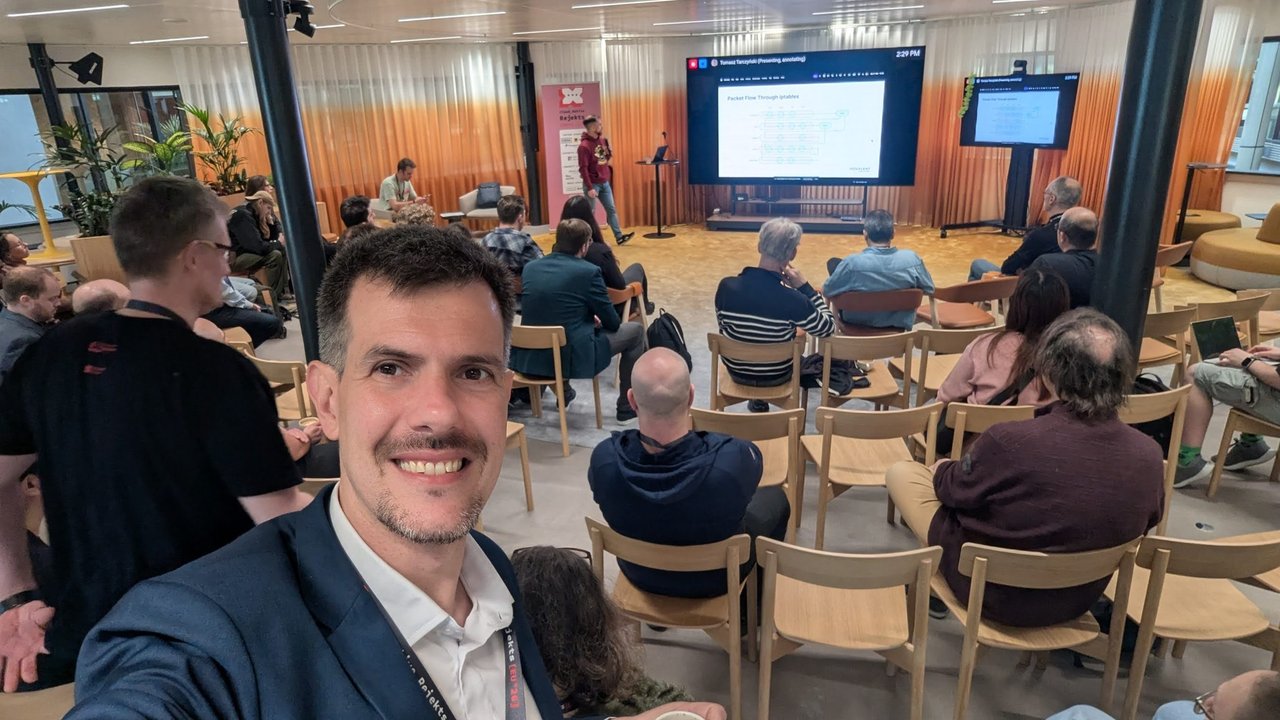 Luca Berton in the audience at Cloud Native Rejekts 2026 while Tomasz TarczyĹski presents a Packet Flow Through iptables slide in Room 2 at Miro Amsterdam