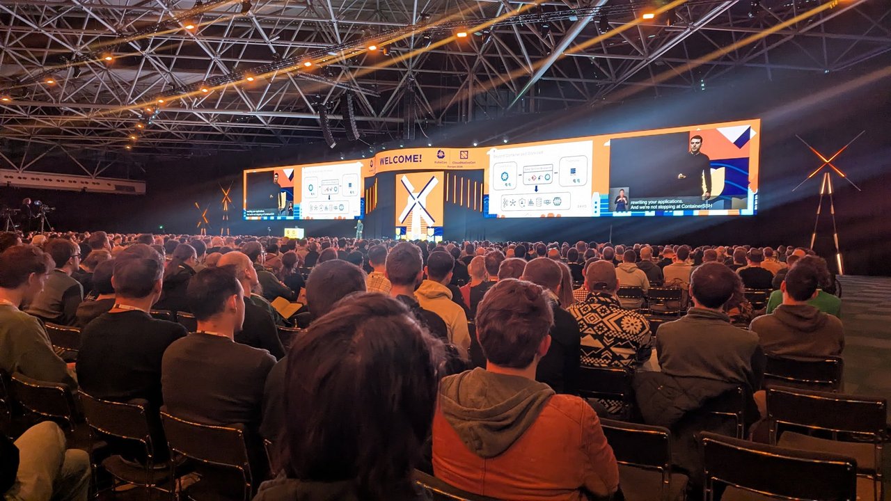 The KubeCon Europe 2026 keynote hall at RAI Amsterdam, with a Beyond Containerized Workload slide from Saxo on the screens and a full audience in front of the stage