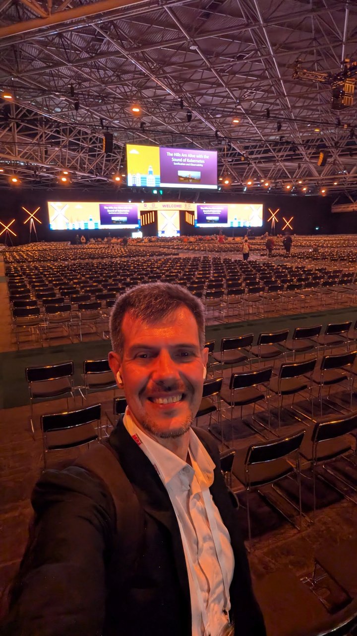 Luca Berton taking a selfie in a large session hall at KubeCon Europe 2026, with the stage and screens lit behind rows of empty seats