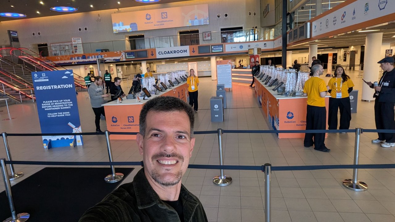 Luca Berton taking a selfie in the KubeCon + CloudNativeCon Europe 2026 registration hall at RAI Amsterdam on Sunday, with the badge desks, a cloakroom sign and a Maintainer Summit registration banner behind him