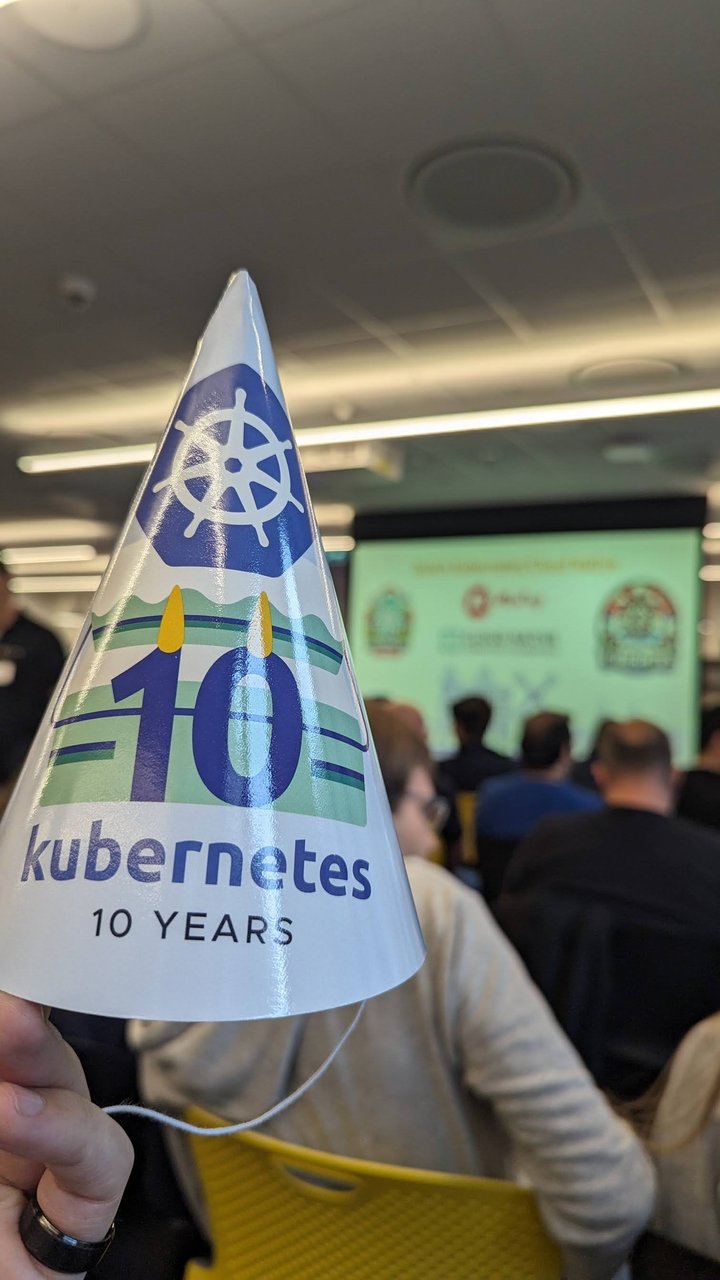 A white party hat printed with the Kubernetes logo and the words kubernetes 10 years, held up in front of the meetup screen