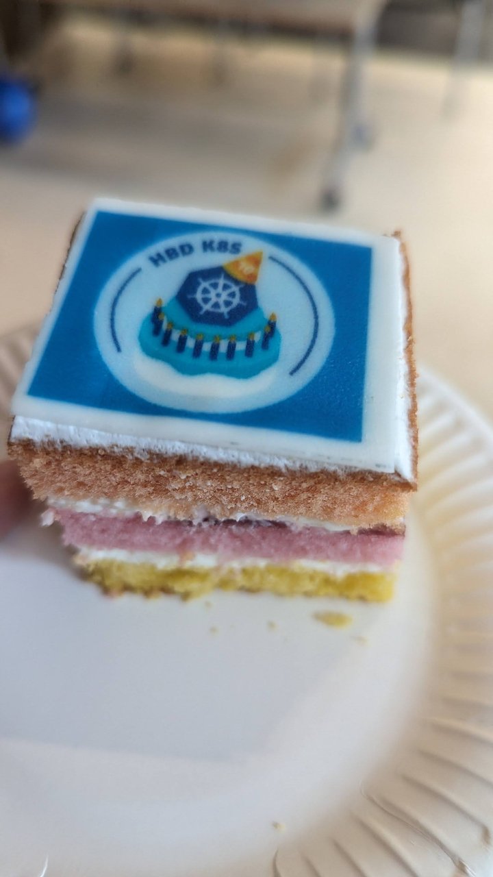 A slice of layered sponge cake with a printed icing picture of the Kubernetes logo and the text HBD K8S