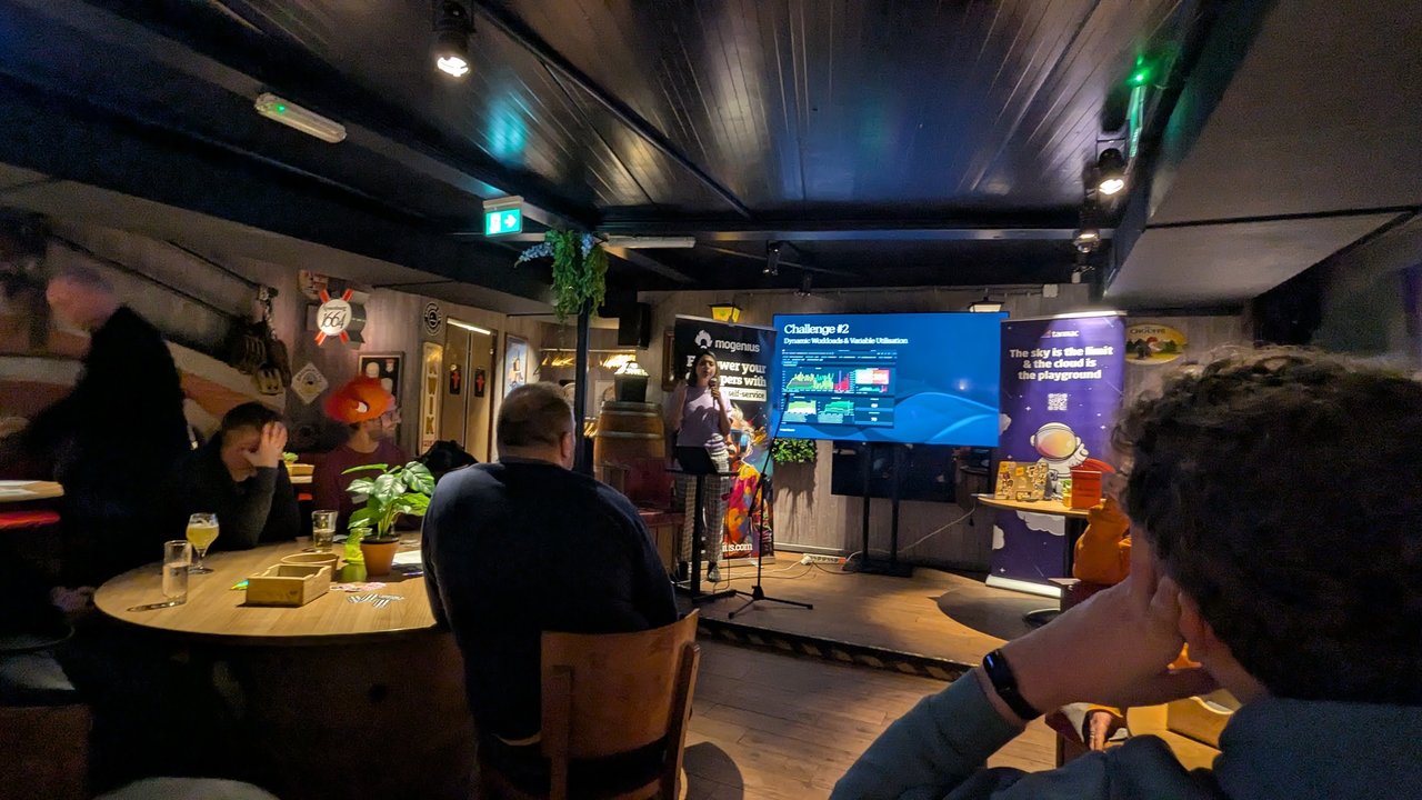 Challenge 2 slide, Dynamic Workloads and Variable Utilisation, showing a monitoring dashboard with spiky usage graphs, in a pub with the speaker at the lectern