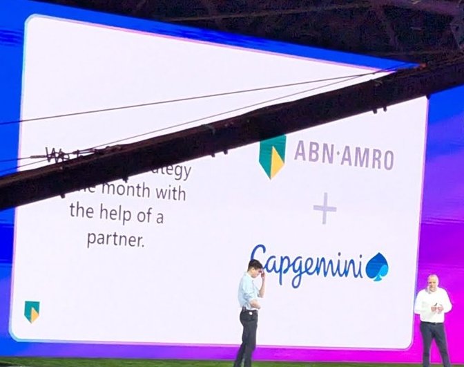 Stage with two speakers in front of a slide showing the ABN AMRO and Capgemini logos and a line about a strategy delivered within a month with the help of a partner, with a large seated audience