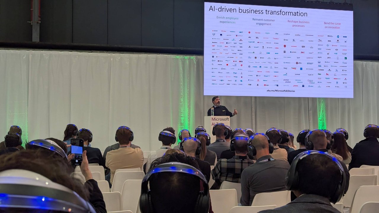 Presenter on a small stage in front of a slide titled AI-driven business transformation with four columns of customer logos, with an audience wearing headphones