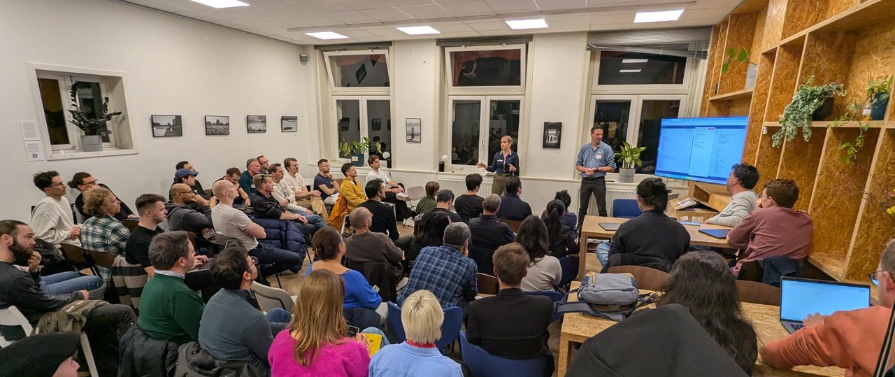 A packed small room at the n8n meetup in Amsterdam with two presenters standing at the front beside a large screen