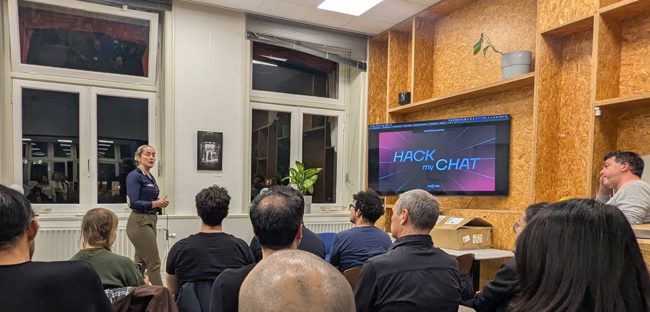 A presenter standing next to a screen showing the slide Hack my Chat in a small room with an audience