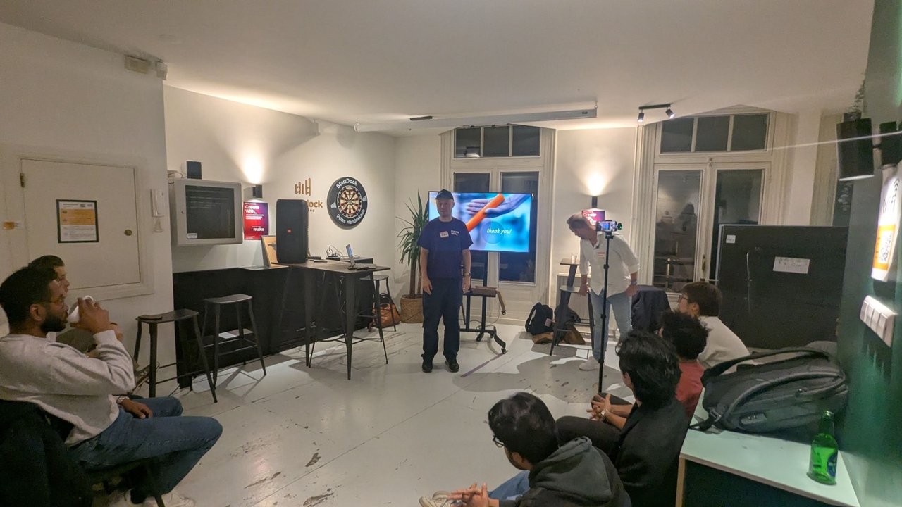 A speaker standing in front of a thank you slide at the end of the evening, with an organiser behind a phone on a tripod