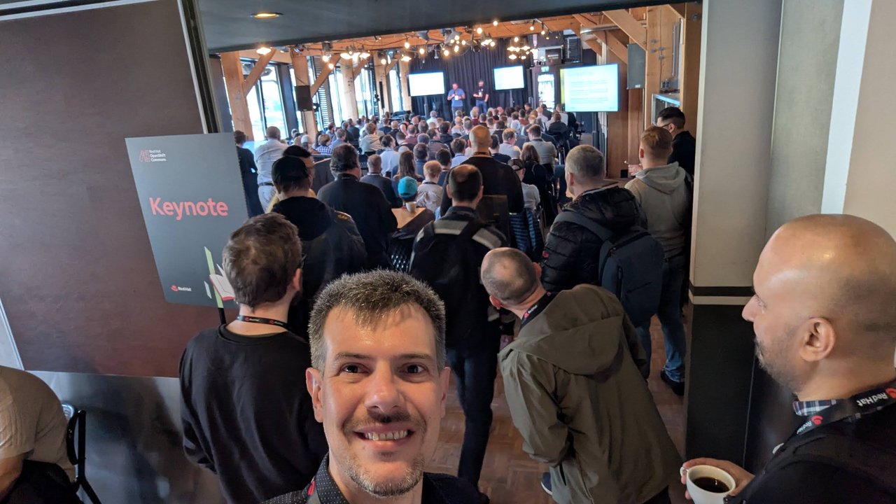 Luca Berton at the Keynote entrance sign at OpenShift Commons Gathering Amsterdam 2026, with a crowd filling the room behind