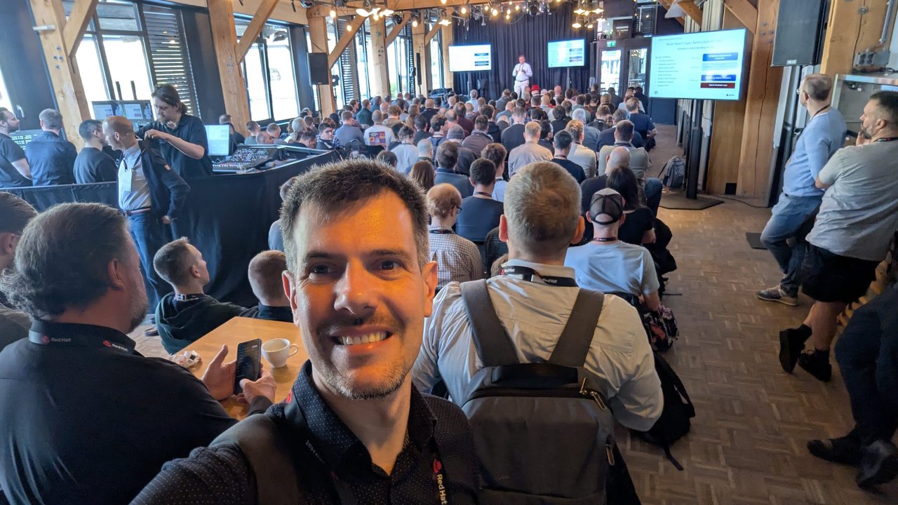 Luca Berton seated in the packed keynote room at OpenShift Commons Gathering Amsterdam 2026, with a speaker on stage and slides on several screens