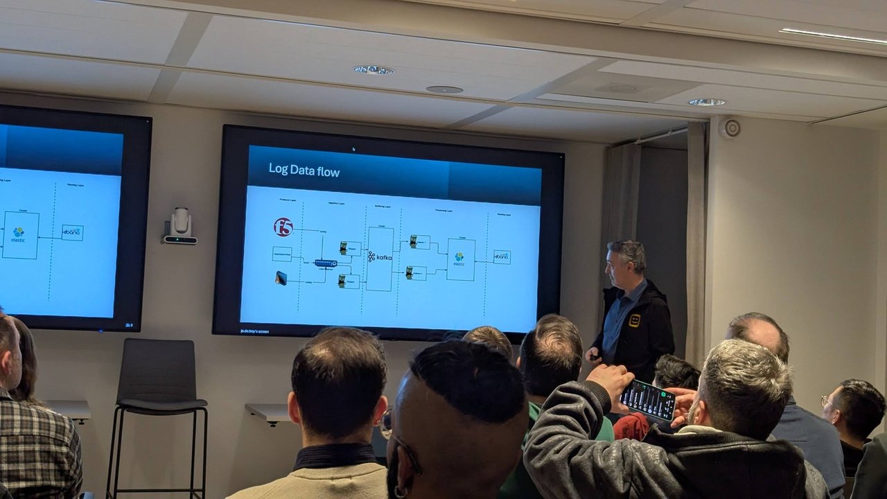 Presentation screen titled Log Data flow showing a Kafka-based pipeline into Elastic and Kibana, with the audience in front
