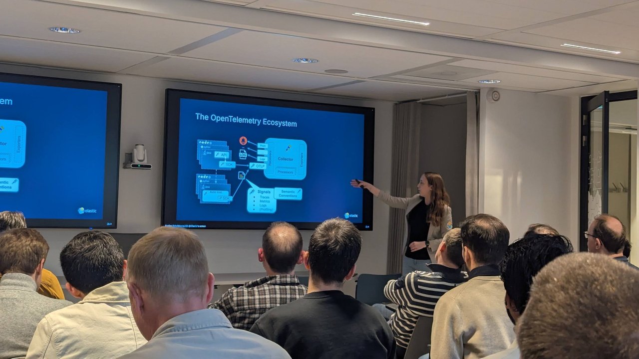 Speaker pointing at The OpenTelemetry Ecosystem slide showing SDKs, OTLP, a Collector with receivers, processors and exporters, and the signals and semantic conventions