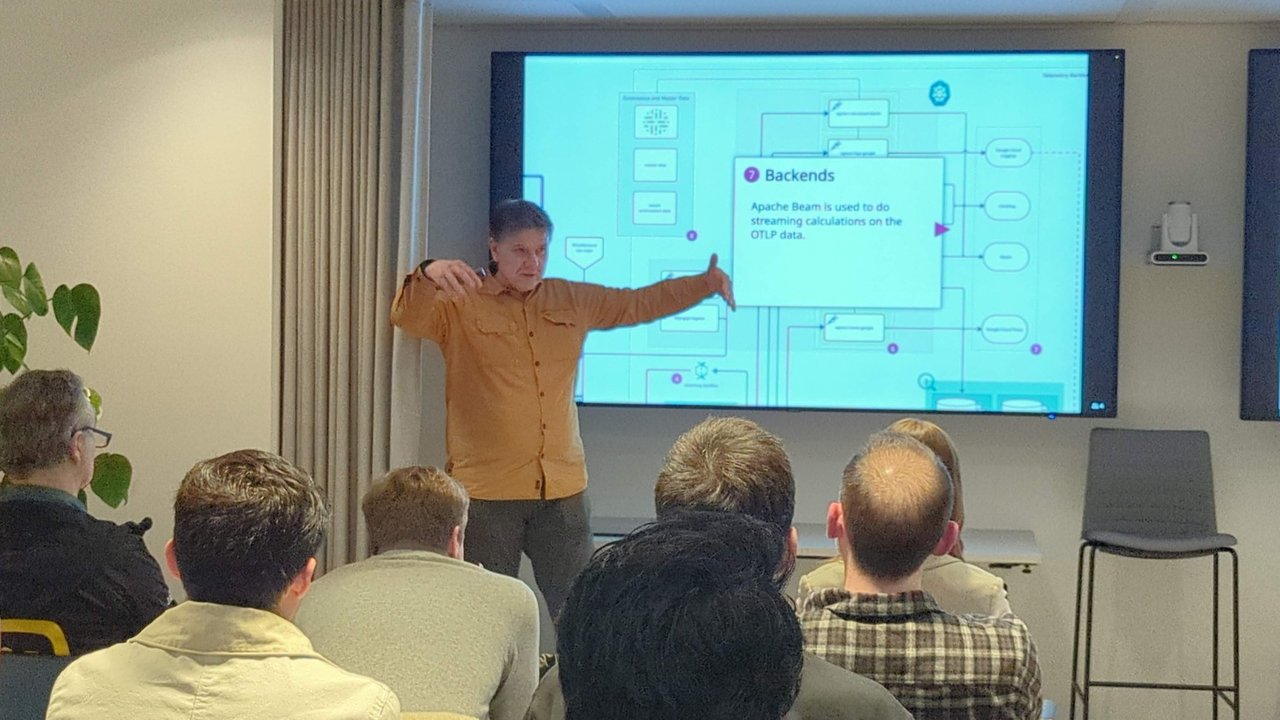 Speaker gesturing at a large architecture diagram with a Backends box saying Apache Beam is used to do streaming calculations on the OTLP data