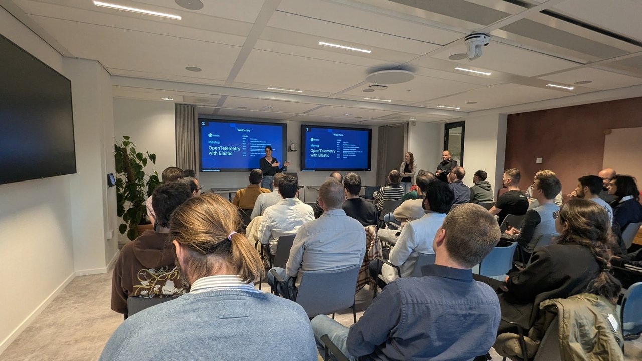 Meetup room with two screens showing the Welcome and agenda slide for the OpenTelemetry with Elastic meetup, two presenters standing at the front and the audience seated