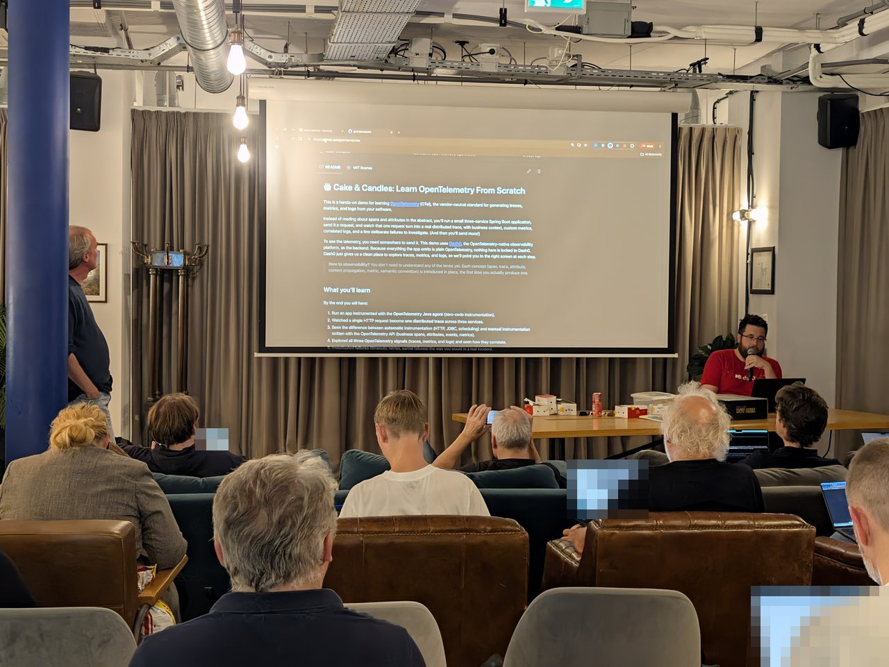 The Cake and Candles README on screen at the OTel meetup, describing a hands-on OpenTelemetry demo with a three-service Spring Boot application, with attendees on laptops