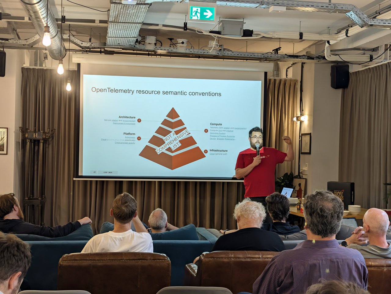 Speaker presenting the OpenTelemetry resource semantic conventions pyramid, with Architecture, Compute, Platform and Infrastructure layers and a Not a comprehensive list banner