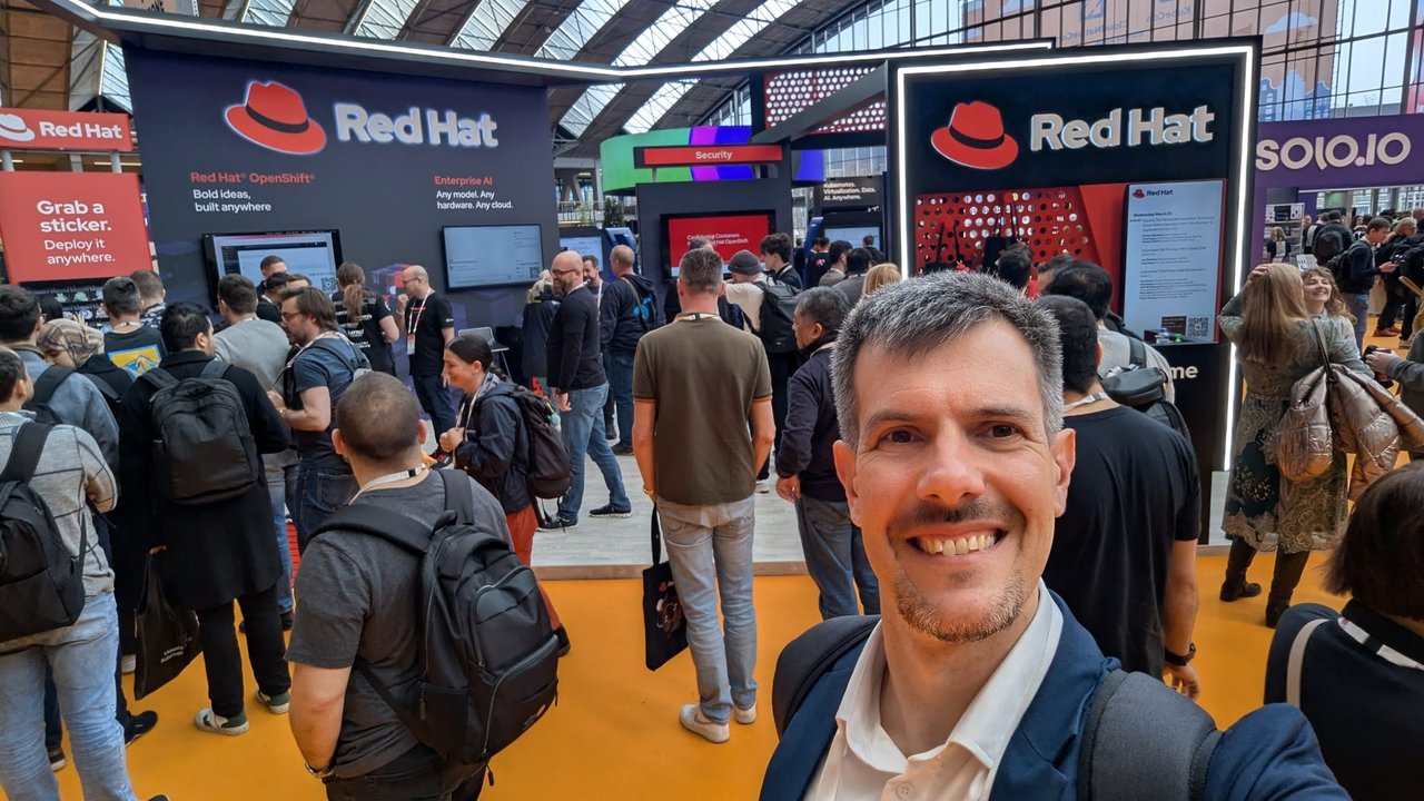 Luca Berton taking a selfie in front of the Red Hat stand on the KubeCon Europe 2026 expo floor, with Red Hat OpenShift and Enterprise AI signs and a crowd of attendees