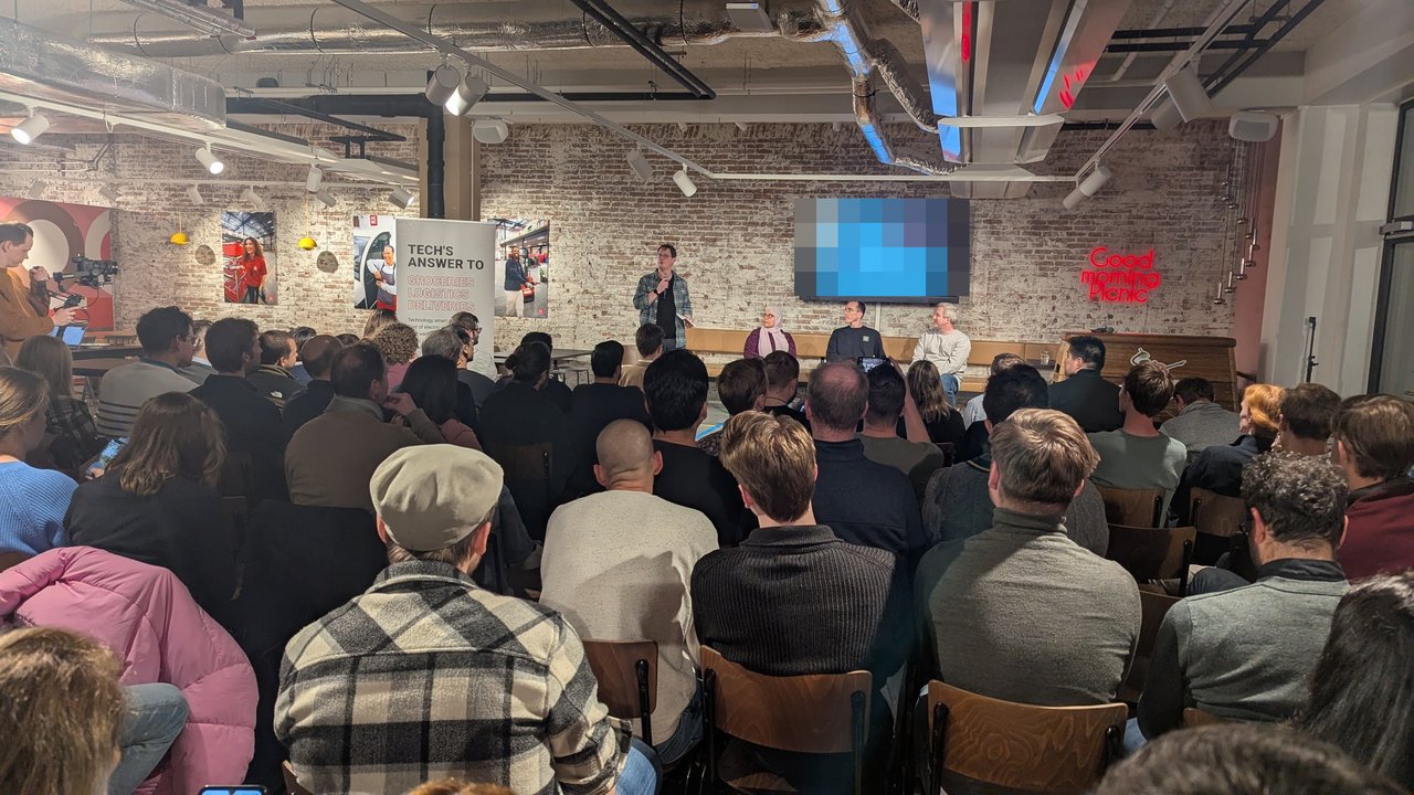 Host opening the developer experience panel at Picnic in Amsterdam in front of a full room, with three panellists seated and the Tech's answer to groceries, logistics, deliveries banner