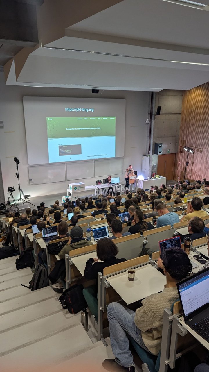 A packed lecture hall at CfgMgmtCamp 2025 in Ghent with the pkl-lang.org homepage on the main screen during the Pkl talk