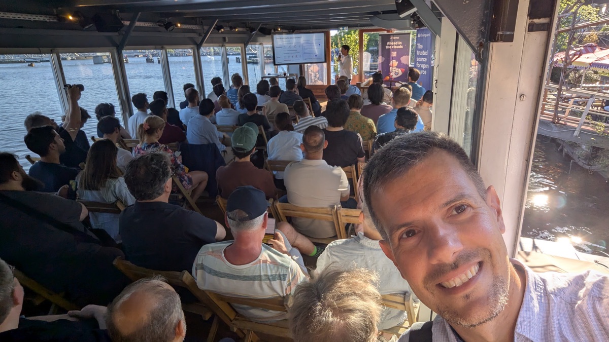 Speaker presenting on a boat at Hannekes Boot with the Amsterdam waterfront in view, Platform Engineering Amsterdam FinTech meetup