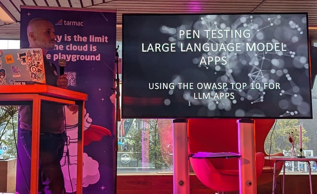 Pen Testing Large Language Model Apps, Using the OWASP Top 10 for LLM Apps title slide next to the tarmac roll-up, with the speaker at the lectern