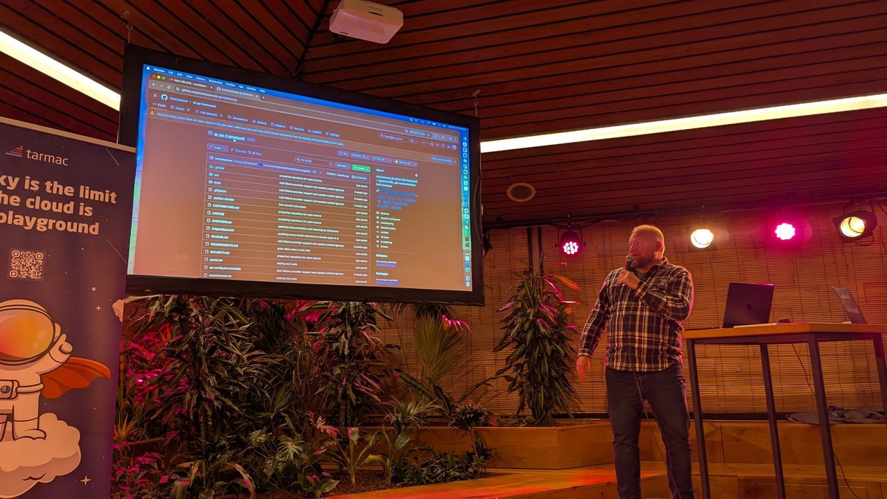 The ai-qa-framework GitHub repository on the big screen at Tolhuistuin, with the presenter holding a microphone beside it and the tarmac roll-up banner on the left